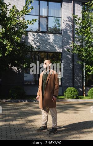 vista completa dell'uomo asiatico in elegante vestito autunnale in piedi vicino edificio e alberi sulla strada Foto Stock