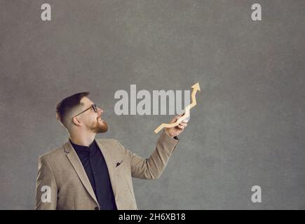 Un uomo d'affari felice tiene una freccia di crescita e la mostra in piedi su uno sfondo grigio. Foto Stock