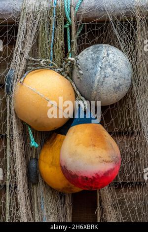 vecchi carri di pesca colorati e boe di plastica appesi alla parete di legno di un capannone di pescatori o capanna con reti che asciugano al sole. Foto Stock