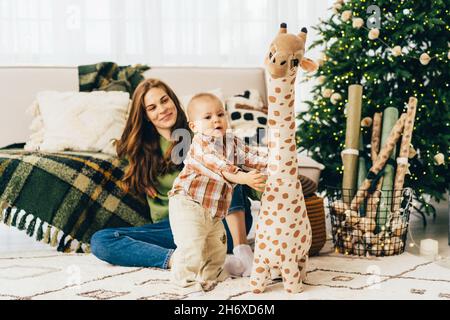 La mummia amorevole si siede sul pavimento e guarda come il bambino gioca con un grande giocattolo di giraffa peluche. Foto Stock