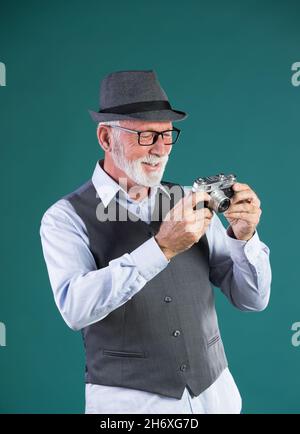 Uomo anziano con barba bianca, in abiti d'epoca e cappello che tiene la vecchia telecamera analogica su sfondo blu in studio Foto Stock