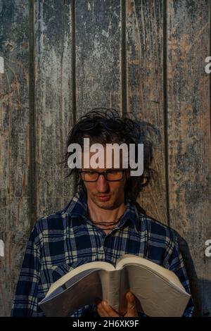 Uomo di capelli neri seduto vicino a una vecchia parete di legno con libro in serata calda al tramonto Foto Stock