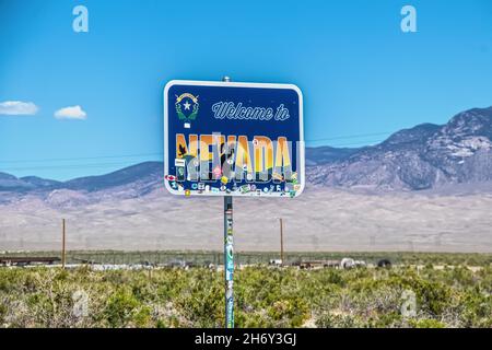 2021 06 Nevada Border USA - Benvenuto in Nevada segno con adesivi parzialmente coprente con filo recinzione e desolato viola montagne sfocate in backgroun Foto Stock