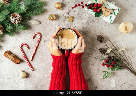 Stesa piatta di mani donna in maglione rosso che tiene una tazza di caffè. Vista dall'alto di sfondo grigio con tazza di bevanda calda, lecca lecca, regalo, ramo di abete, pino Foto Stock