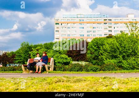 POZNAN, POLONIA - 16 maggio 2014: Due donne sedute su una panca di legno da un sentiero in un parco vicino ad un alto edificio di appartamenti a Poznan, Polonia Foto Stock