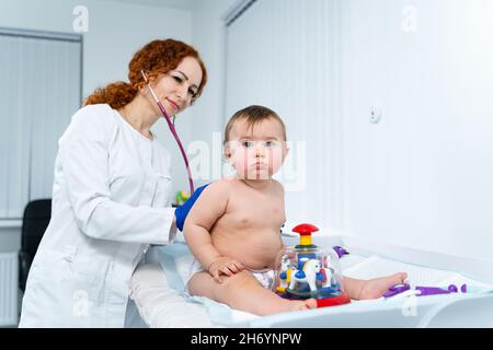 Il medico femminile dai capelli rossi esamina il bambino piccolo per meno di un anno in una clinica moderna. Pediatra durante l'esame del bambino in ospedale Foto Stock