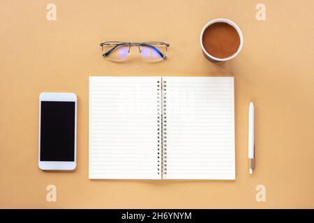 Blocco note vuoto, penna, caffè in una tazza di carta, smartphone e bicchieri su tavolo beige. Vista dall'alto, piatto, mock up. Foto Stock