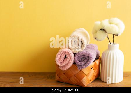 Pila di asciugamani da bagno in cestino e pianta di cotone su tavola di legno. Sfondo giallo. Spazio copia Foto Stock