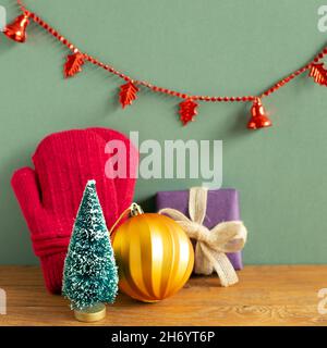 Inverno natale concetto. Guanti invernali, scatola regalo, albero, bauble, garland partito su tavola di legno. Sfondo verde. Spazio copia Foto Stock