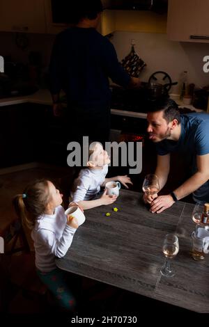 L'uomo mostra la lingua ai bambini, i bambini anche la lingua. Famiglia party amici a casa. Un uomo cucina il cibo. Adulti bere vino. Foto Stock