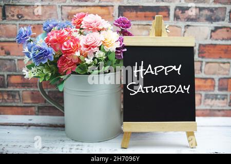 Testo del Sabato felice su cavalletto in legno nero e vaso con bouquet di fiori su sfondo muro di mattoni Foto Stock