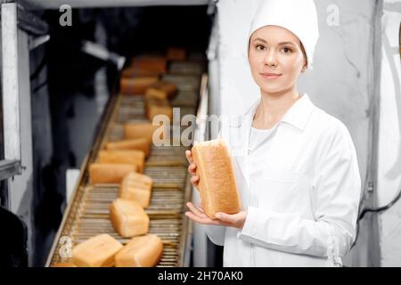 Baker donna che tiene il pane fresco in mani sul trasportatore automatico di fondo della fabbrica di panetteria alimentare. Foto Stock