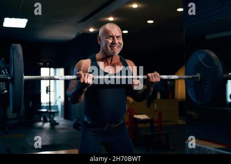 Emotivo più vecchio Bodybuilder bald treni in palestra, mentre pompando i muscoli bicipite con barbell Foto Stock