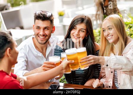 Gruppo di amici positivi in abiti casual seduti al tavolo e calici di birra mentre festeggia il compleanno al bar durante il giorno Foto Stock
