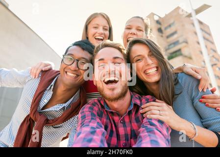 Ritratto ad angolo basso di un allegro gruppo interrazziale di cinque amici sorridenti mentre si scattano foto selfie all'aperto in città Foto Stock