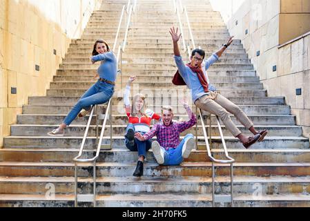 Un gruppo multiculturale di amici felici ed eccitati che si acclamano e ridono mentre scivolano sulle rotaie di una rampa di scale esterne in una città in libertà Foto Stock