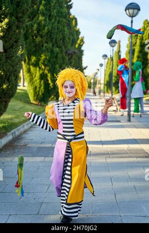 Felice clown femminile con volto dipinto in costume divertente e parrucca gialla, sorridente e camminando sul sentiero nel parco con poi colorato nelle mani durante la sfilata Foto Stock