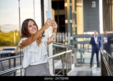 Vista laterale della giovane donna in abiti casual con selfie su smartphone mentre si trova sul ponte della città Foto Stock
