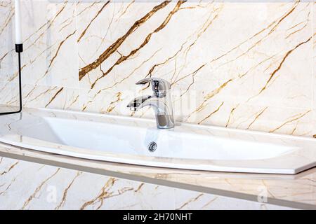 Nuovo e moderno rubinetto d'acqua in metallo con lavandino bianco sullo sfondo della parete in marmo nella stanza da bagno. Foto Stock