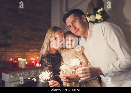 Famiglia felice - madre, padre e figlia che tiene scintille sullo sfondo di un albero di Natale e camino con candele. Foto Stock