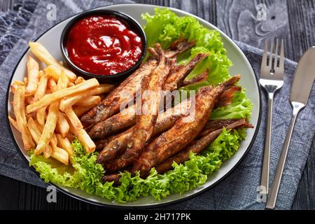 pesce fritto croccante di capelin con lattuga fresca, patatine fritte e salsa di pomodoro su un piatto su un tavolo di legno nero testurizzato Foto Stock