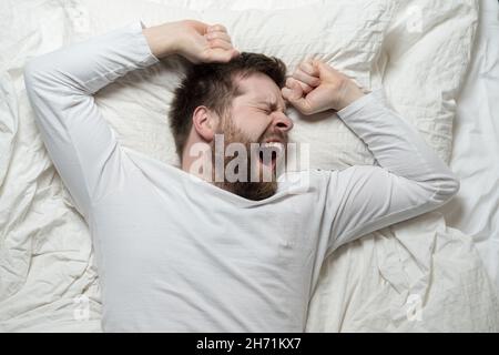 L'uomo bearded in pigiama bianca è stanco e vuole dormire, lui grida e si allunga mentre si trova a letto. Vista dall'alto. Primo piano. Foto Stock