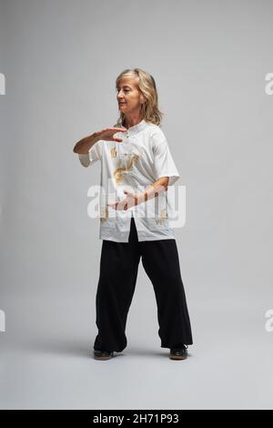 Donna bionda matura praticando Chi Kung e Tai Chi su sfondo bianco. Indossa una tradizionale giacca cinese Tai Chi bianca, pantaloni neri e bl Foto Stock