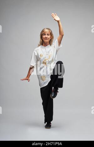 Donna bionda matura praticando Chi Kung e Tai Chi su sfondo bianco. Indossa una tradizionale giacca cinese Tai Chi bianca, pantaloni neri e bl Foto Stock