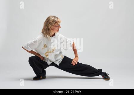 Donna bionda matura praticando Chi Kung e Tai Chi su sfondo bianco. Indossa una tradizionale giacca cinese Tai Chi bianca, pantaloni neri e bl Foto Stock