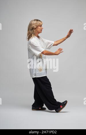 Donna bionda matura praticando Chi Kung e Tai Chi su sfondo bianco. Indossa una tradizionale giacca cinese Tai Chi bianca, pantaloni neri e bl Foto Stock