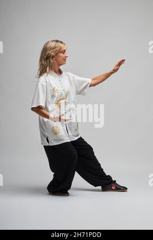 Donna bionda matura praticando Chi Kung e Tai Chi su sfondo bianco. Indossa una tradizionale giacca cinese Tai Chi bianca, pantaloni neri e bl Foto Stock