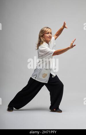 Donna bionda matura praticando Chi Kung e Tai Chi su sfondo bianco. Indossa una tradizionale giacca cinese Tai Chi bianca, pantaloni neri e bl Foto Stock