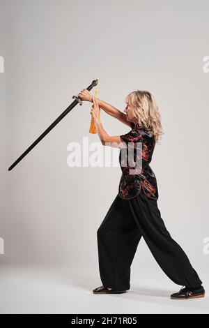 Donna bionda matura praticando Tai Chin con spada su sfondo bianco indossando una tradizionale giacca cinese nera con decorazione cinese rossa, nera Foto Stock