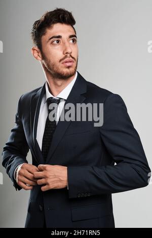 Giovane uomo con barba che indossa un abito blu scuro. Sta fissando la giacca del vestito mentre guarda alla sua sinistra. Foto Stock