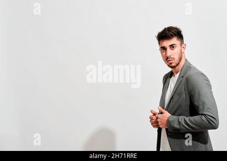 Giovane uomo che indossa jeans, una tshit bianca e un blazer grigio che cammina di fronte a una parete bianca e guarda la macchina fotografica. Foto Stock