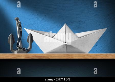 Primo piano di una barca di carta bianca e di un ancoraggio di metallo grigio con catena su una mensola di legno a casa, con una parete blu su sfondo. Foto Stock