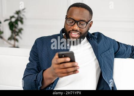 Primo piano ritratto di uomo afro-americano millenario in occhiali utilizzando smartphone per trascorrere il tempo libero, incontri, chat in social network, ragazzo nero allegro guardando sullo schermo del telefono Foto Stock