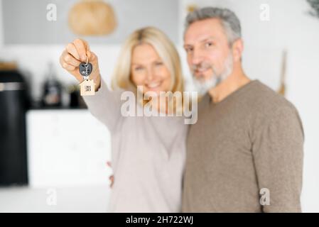 Fuoco selettivo sulle chiavi con portachiavi in forma di piccola casa in mano femminile, allegro coppia di mezza età tiene le chiavi dalla nuova proprietà, acquirenti felici di una propria proprietà. Coniugi ottenere un mutuo Foto Stock