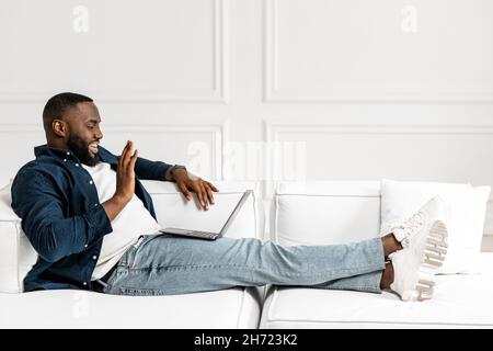 L'uomo africano di buon aspetto sta usando un laptop per la connessione video, facendo le chiamate reali, la riunione attesa da tempo, diciamo ciao, guardando lo schermo del calcolatore, disteso sul divano e tenendo il laptop elegante sul grembo Foto Stock
