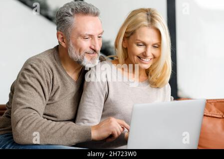 I coniugi maturi adorabili utilizzano un computer portatile per trascorrere il tempo libero a casa, guardare film insieme, scegliere acquisti, fare shopping. La coppia di mezza età si siede sul divano e guarda sullo schermo Foto Stock
