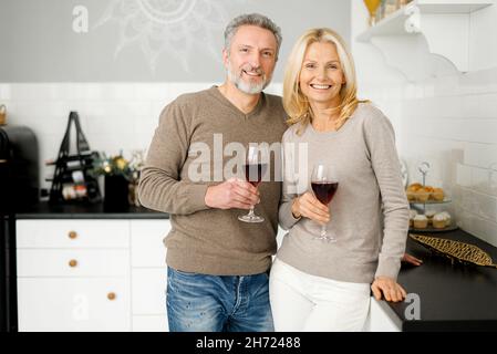 Coppia di mezza età che tiene bicchieri di vino guardare la macchina fotografica e sorrisi in piedi in cucina a casa, uomo maturo grigio-capelli e affascinante donna di mezza età celebrare l'anniversario, hanno data Foto Stock