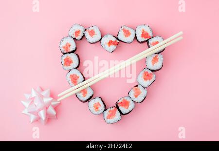 Sushi di salmone rotolini disposti a forma di cuore su sfondo rosa, con bacchette a forma di freccia Cupido, prua vacanza. Concetto - Valenti Foto Stock