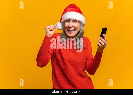 Sorpresa felice donna anziana in santa Hat tenendo smartphone e gioisce, la donna di mezza età tiene un telefono, ha ricevuto un messaggio o una notifica inaspettata, guarda la fotocamera stupito, dice sì, isolato Foto Stock