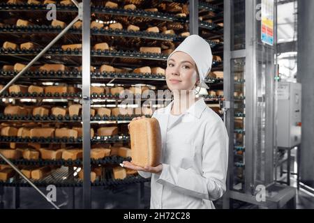 Baker donna che tiene il pane fresco in mani sul trasportatore automatico di fondo della fabbrica di panetteria alimentare. Foto Stock