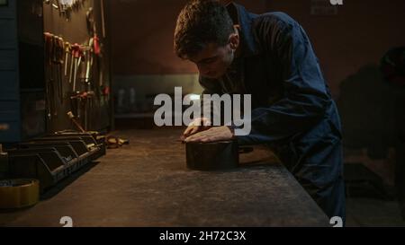 Giovane uomo fabbro che leviga un pezzo di ferro o di acciaio nella sua officina Foto Stock