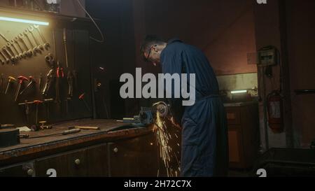 Il giovane uomo fabbro usa la macchina da taglio del ferro o del metallo e la macinazione del metallo sul pezzo di ricambio dell'acciaio in officina Foto Stock