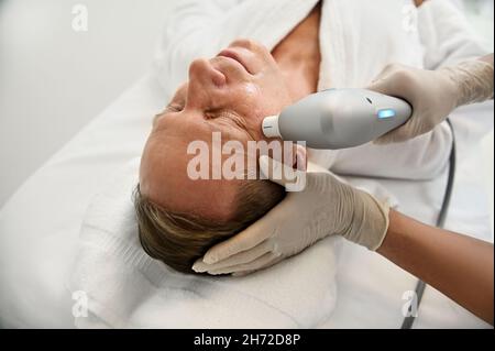 Primo piano. Mano estetista che tiene moderno apparato medico ultrasonico e che effettua il sollevamento del viso e ringiovanente trattamento di bellezza anti-invecchiamento su fa Foto Stock