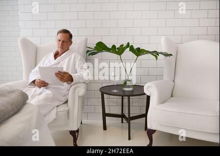 uomo d'affari di 50 anni con accappatoio bianco spugna leggere listino prezzi catalogo delle procedure di bellezza seduta su una poltrona di una lussuosa wel bianca Foto Stock