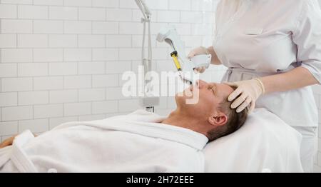 Bell'uomo anziano che indossa un vestito di spugna bianca, sdraiato sul tavolo da massaggio e che riceve l'iniezione di bellezza facciale con le moderne attrezzature in wellnes di lusso bianche Foto Stock