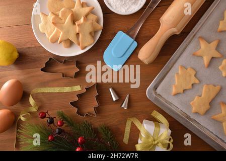 Tavolo in legno con biscotti con forme natalizie appena sfornate su una teglia da forno e piatto con stampi e utensili da cucina. Vista dall'alto. Composizione orizzontale Foto Stock